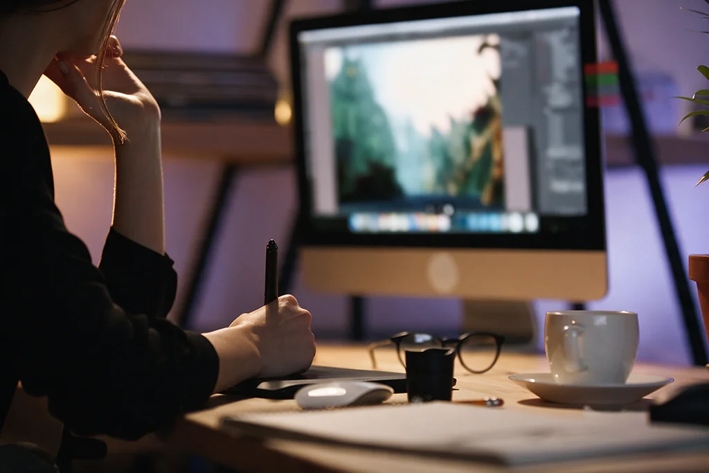 Persona usando el lápiz de una tableta gráfica mientras edita la foto de un bosque en un monitor de escritorio, junto a unas gafas, una taza y un ratón