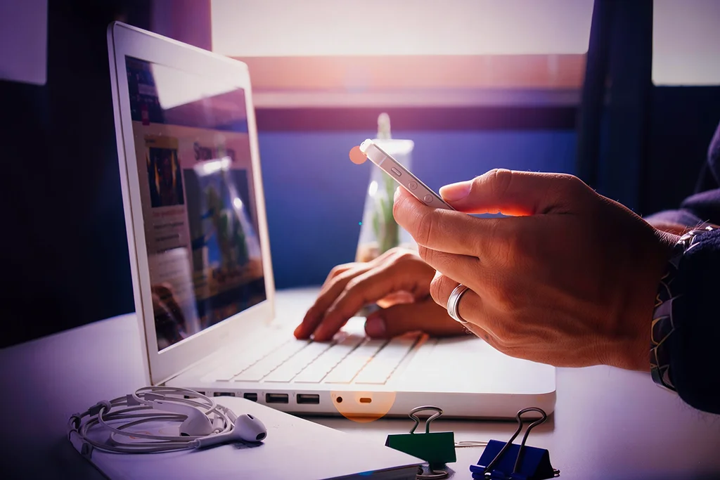 Persona revisando una página web en un portátil mientras sostiene un teléfono móvil con la otra mano, en un escritorio con auriculares y pinzas de papel