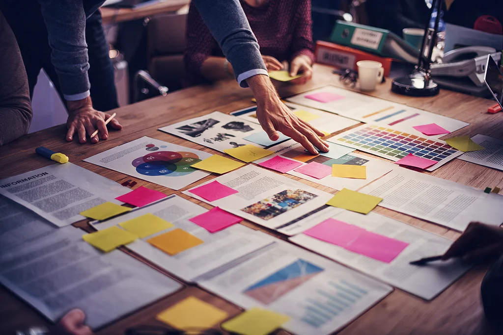 Varias personas alrededor de una mesa con documentos impresos, una carta de colores, gráficos y notas adhesivas rosas y amarillas; una mano señala uno de los papeles